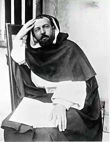 Photo noir et blanc d'un homme en toge de prédicateur (toge noire sur toge blanche) d'une quarantaine d'années. Sa tête est dégranie sur le devant, il a les cheveux noirs et la barbe noire. Il est assis confortablement sur une chaise posant la main droite sur la tempe droite et la main gauche sur un livre ouvert posé sur ses genoux. L'arrière plan est constitué d'un mur blans et d'une fenê pourvue de barreaux.