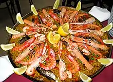 Photographie couleur représentant un plat garni. Le riz safrané garni de petits pois et de mollusques et surmonté de langoustines, moules et quartiers de citron.