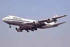 Photo du côté gauche, légèrement de face, d'un avion en vol aux couleurs blanches et bleues de Pan Am avec le train d'atterrissage sorti.