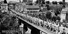 Photo en noir et blanc montrant un peloton de cycliste très étendu passant sur un pont avec de nombreux spectateurs.