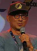 Photo en buste d'un homme âgé portant des lunettes et une casquette et tenant un micro à la main.