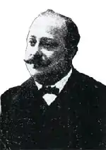 Portrait photo de mauvaise qualité d'un homme 50-60 ans, front dégarni et moustache, costume sombre et nœud papillon.