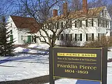 Photographie d'une maison blanche à deux étages avec de hautes cheminées en brique. Le sol est recouvert de neige et un panneau indique qu'il s'agit de la résidence du 14e président des États-Unis.