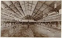 Vue du grand bassin de la piscine avec des nageurs sous un toit du type halle. Au fond du bassin sur le petit côté se trouvent des gradins.