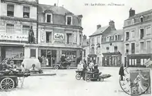 Carte poste en noir et blanc présentant la place Henri IV au début du XXe siècle : des marchandes des quatre-saisons devant la statue, les raisons sociales des magasins peintes sur les murs, un timbre oblitéré en bas à droite.