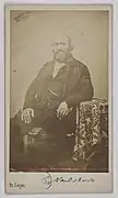 Photographie en noir et blanc représentant un homme mûr barbu en costume trois pièces sombre assis de trois-quart face sur un fauteuil et accoudé à un guéridon recouvert d'un tissu.