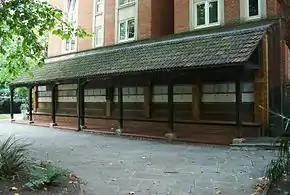 Une longue structure sombre en bois avec un auvent abritant un mur portant des rangées parallèle de plaques de céramiques de couleur claire.