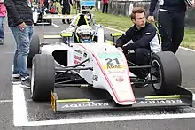 Photographie d'une Formule 4 blanche et jaune, vue de face, à l'arrêt sur une grille de départ. Le pilote est dans la voiture, entouré de deux mécaniciens.