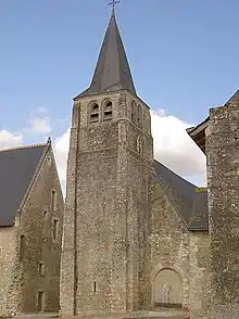 Photographie en couleurs d'un clocher à base carrée et étages crénelés surmontés d'une flèche octogonale recouverte d'ardoises, deux bâtiments venant l'encadrer de part et d'autre, une nef munie d'une porte cintrée visible en arrière-plan.