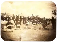 Une vieille photo montrant une procession passant entre des rangées de soldats avec des tentes en toile de fond