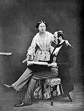 Photographie en noir et blanc d'un couple. L'homme est assis et lit un livre. La femme se tient derrière lui.