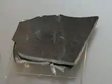 Tesson de ustensile de vaissellerie en céramique de type bucchero nero datant du 1er&nbsp;quart du VIe&nbsp;siècle&nbsp;av. J.-C. L'artéfact de terre cuite porte le graphe écrit en caractères étrusques :  (autrement dit, « Rex »),,,