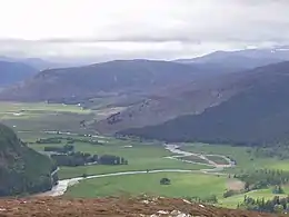 Photo. Vallée large et verdoyante avec rivière, entourée de montagnes au ton violet et en partie couvertes de bruyère.