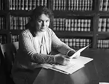 Une jeune femme à la peau claire et aux cheveux noirs, assise à une table, tenant des papiers, devant une bibliothèque remplie d'ouvrages juridiques.
