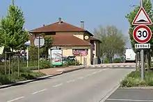 Une route avec deux panneaux à droite dont un indique que la voie est limitée à 30 km/h. L'autre panneau indique la présence d'un ralentisseur. À gauche de la route se trouve un abri-bus devant un bâtiment jaune faisant office de restaurant.