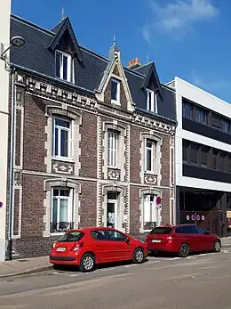 Cliché numérique couleur. Devant une maison de la fin du XIXe siècle et un immeuble du XXe siècle de trois étages stationnent deux voitures rouges.