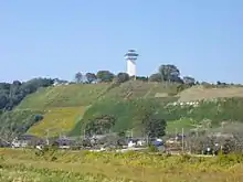 Photo couleur d'une colline couverte de verdure, avec un village à son pied, et une tour blanche à son sommet, sur fond de ciel bleu.