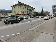 Photographie en couleurs d’un arrêt de bus en zone urbaine, avec plusieurs voitures roulant à proximité et des maisons.