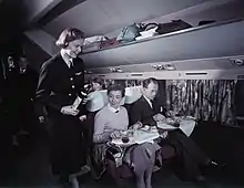 passagers installés, une hôtesse de l'air en uniforme noir leur présente une bouteille de vin.