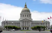 Un grand bâtiment sur trois étages avec une aile de chaque côté d'une tour imposante. Le bâtiment est blanc, avec un dome or et bleu au sommet de la tour. Devant le bâtiment on trouve une place encadrée de mâts pour drapeau et d'arbres.