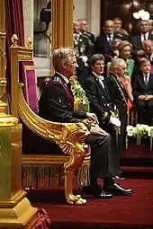Photographie de profil d'un homme aux cheveux blancs assis sur un fauteuil aux accoudoirs dorés
