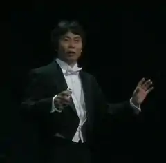 Photo d'un Japonais en costume de cérémonie noir sur scène de fond noir également.