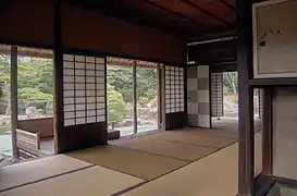 Grande salle, pour le repas, cuisine extérieure ; arrière-plan : petite salle pour le thé. Poteaux peu équarris.