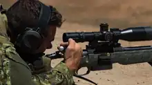 Photographie couleur. Visage de profil orienté vers la droite d'un militaire équipé d'un casque anti-bruit et épaulant, allongé, avec un fusil à lunette. De sa main droite, l'homme tire la culasse de son arme et en éjecte une douille