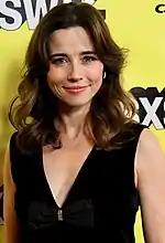 Femme brune souriante en robe noire devant une affiche au fond jaune portant les lettres SXSW du festival South by Southwest.