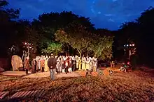 Répétition à la nuit tombante d'une cinquantaine de personnes en costumes, dont quelques enfants. La scène; éclairée par de projecteurs, est dans une clairière entourée d'arbres