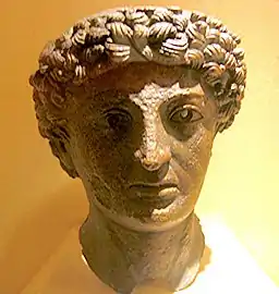 Tête en bronze vert, avec les cheveux bouclés formant une grosse couronne, à demi plongée dans l'ombre.