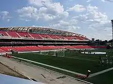 Un terrain de sport vert, des gradins rouges et un ciel bleu comprenant quelques nuages.