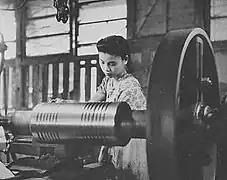 Photo d'une ouvrière en costume traditionnel travaillant sur une machine, visible au premier plan.