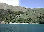 Photographie d'un littoral très pentu et déboisé en partie haute, au pied duquel quelques maisons et un ponton sont visibles.