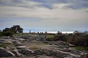 Photo d'un plateau rocheux avec de la végétation et des gens au fond. Au second plan la mer, les montagnes et le ciel.
