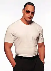 Un catcheur musclé au t-shirt blanc et au pantalon noir avec des lunettes de soleil pose devant un fond blanc avec ses mains dans les poches.