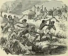 Plusieurs groupes de personnes noires se battent dans les hautes herbes avec des armes. Dans l'arrière plan de l'image se trouve un village avec des activités festives et ce qui ressemble à un homme pratiquant à un rituel.