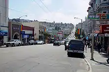 Une rue se développe jusqu'à l'arrière-plan. Des magasins sont présents des deux côtés de la rue, et de nombreux réverbères arborent les drapeaux arc-en-ciel.
