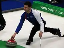 un curleur en pantalon noir, sweater bicolore blanc et bleu s’apprête à lâcher sa pierre de la main droite : large fente des jambes, bras gauche en appui sur son balai, en arrière plan marquage Vancouver 2010 en blanc sur vert