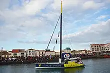 Un voilier bleu marine et jaune descend un chenal au moteur. Sur la coque, un colibri et les inscriptions « Le Projet Imagine » et « Le Souffle du Nord ». Une foule est massée sur le quai.