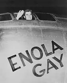 Photographie d'un homme saluant depuis le hublot du cockpit d'un avion sur lequel est écrit "Enola Gay"
