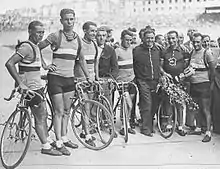 Photographie en noir et blanc d'un groupe de cyclistes se tenant debout à côté de leurs vélos à l'arrivée d'une course.
