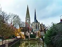 Paysage d'hiver, arbres partiellement dénudés, avec une église gothique, chœur avec arcs-boutants toit en ardoise, surmontée d'une flèche pointue recouverte aussi d'ardoises, et un clocher conique en pierre ; au premier plan un bassin d'eau verte.