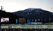 Une foule multicolore regarde passer un bobsleigh noir et blanc sur une piste de bobsleigh marquée des anneaux olympique et de grandes lettres «&nbsp;vancouver 2010&nbsp;» ; derrière, se trouve un écran géant vidéo retransmettant la scène, un autre écran détaillant des résultats sportifs, une rangée de bannières olympiques et de plusieurs pays ; encore plus loin, un paysage de petite montagne partiellement enneigée, et un petit dirigeable d'observation dans le ciel