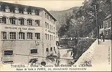 Vieille photographie de l'usine prise avec une toile pendue à une fenêtre du 1er étage où un premier diable rouge est peint au début du XXe&nbsp;siècle.