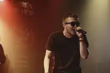 Un homme rappe dans un micro, il porte des lunettes de soleil.