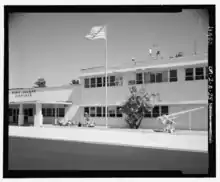 Photographie en noir et blanc du terminal de l'aéroport de l'île Wake, le drapeau américain est hissé devant. Un canon se situe à la droite de l'image. Le tout est entouré de noir.