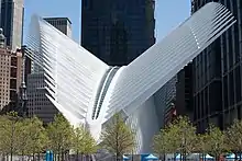 Une structure blanche courbée avec des côtes saillantes formant un angle, vue avec des arbres en herbe devant et des tours modernes derrière elle au soleil avec un ciel bleu clair derrière