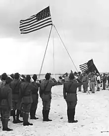 Photographie en noir et blanc représentant le hissage du drapeau américain au milieu de l'image. L'image est divisé horizontalement en deux plans représentant le sable, l'océan et des soldats américains à droite et japonais à gauche ; le second représente l'océan et le drapeau. Sur la partie gauche, un autre drapeau américain flotte au vent.