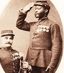 Photographie noir et blanc de deux soldats décorés en uniforme. Un, de couleur blanche, est assis et regarde de face. L'autre, de couleur noire, effectue le salut militaire.